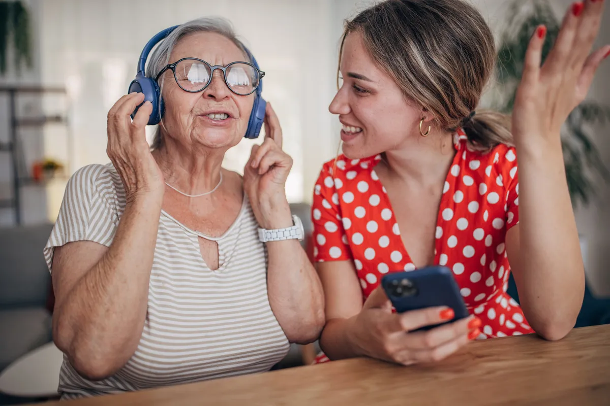 Senior woman with dementia listening to personalized music through headphones, smiling with a loved one.