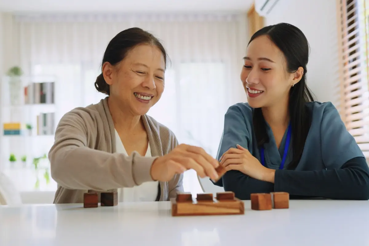 A caregiver in blue scrubs sits with an elderly woman at Carefield Madonna Gardens, engaging in an activity as part of occupational therapy interventions for dementia.