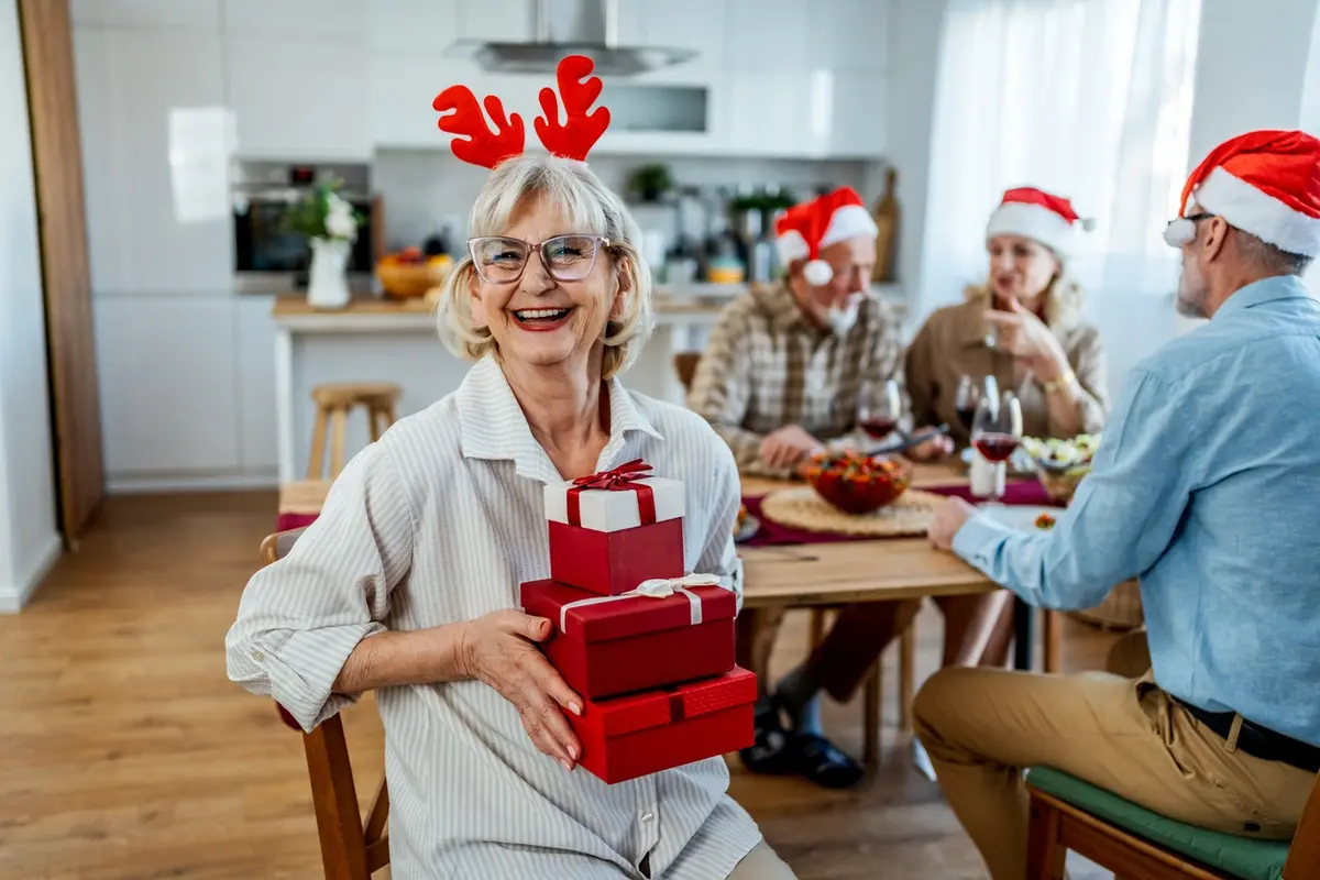 Smiling senior woman holding wrapped Christmas gifts while celebrating with friends in a cozy holidays to residents.