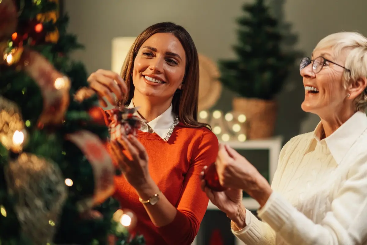 first-holiday-in-senior-living-ojai-christmas-tree Adult daughter helping her senior mother decorate a Christmas tree in a cozy living room. Her first holiday in senior living.