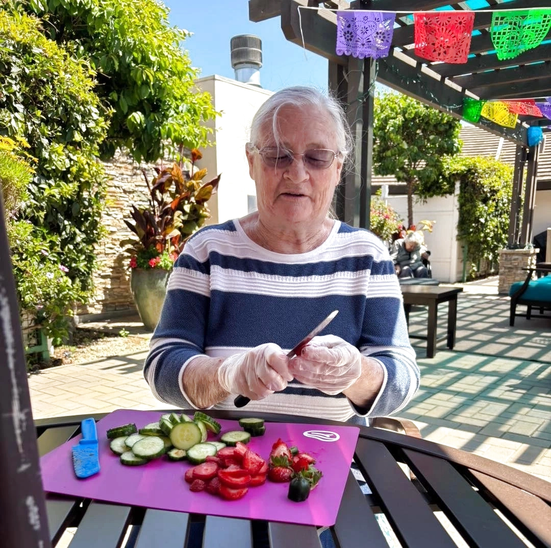 Outdoor Spaces in Assisted and Memory Care Patio Food Activity Outdoor spaces in assisted and memory care: resident slices vegetables on a sunny patio with care.