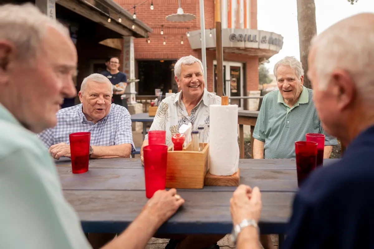 Male residents laughing and socializing at an outdoor dining table during an assisted living Silver mens club outing.