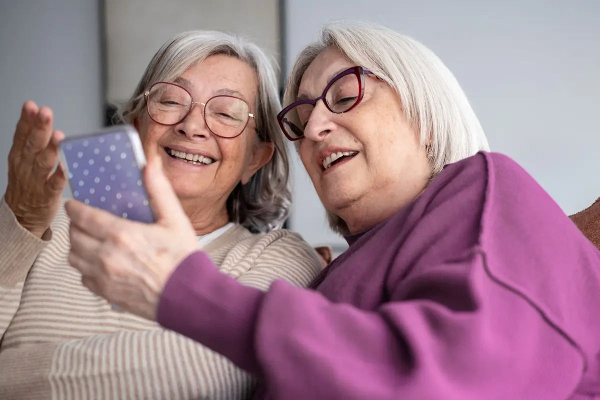 memory-care-companion-suites-pleasanton Two senior women laughing and looking at a smartphone together in a memory care companion suites.